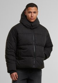 Svart polstret jakke med høy, strukturert krage, frontglidelås og fremtredende quiltede seksjoner. Stoffet virker jevnt med en matt overflate.