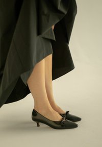 Scarpe nere in vernice con tacco basso, punta affusolata e dettaglio a fiocco. Accompagnate da un vestito nero fluente che arriva poco sopra le caviglie.
