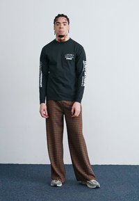 Long-sleeve black shirt with white graphics on sleeves, paired with brown plaid wide-leg trousers and grey sneakers, standing against a plain backdrop.