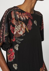 Svart kortärmad topp med intrikat blommönster i röd och beige, med texturerade accenter längs axlarna.