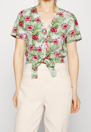 Frau trägt eine kurzärmelige, vorne gebundene Bluse mit roten Blumen und grünen Blättern, kombiniert mit hoch taillierten hellbeigen Hosen.