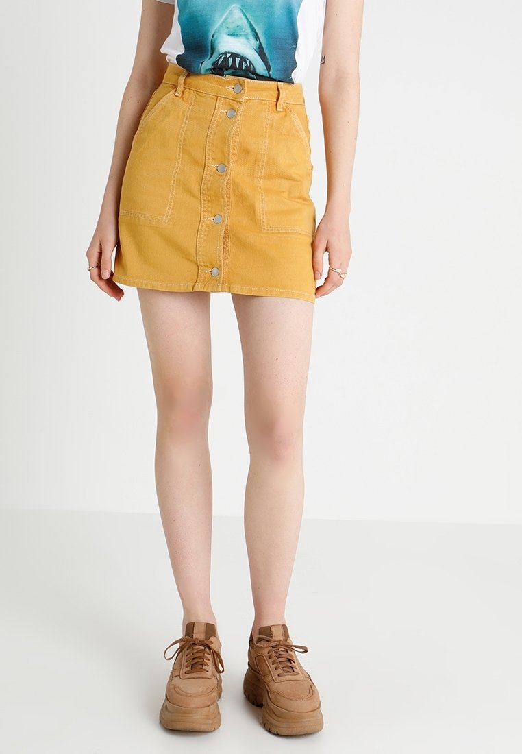 Jupe mini en denim jaune avec fermeture à boutons sur le devant, deux poches avant, et détails de surpiqûres. Associée à des baskets épaisses marron.