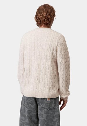 Persoon met golvend bruin haar die een crème kabelgebreide trui en donkergrijze spijkerbroek met patroon draagt, met de rug naar de camera tegen een effen achtergrond.