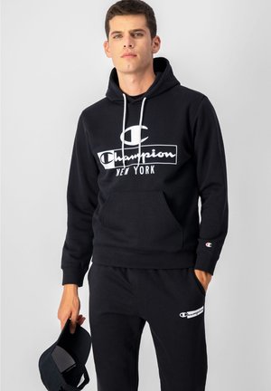 Felpa nera in cotone con cappuccio e coulisse, caratterizzata da un logo Champion bianco e il testo "NEW YORK". Abbinata a pantaloni di felpa neri coordinati.