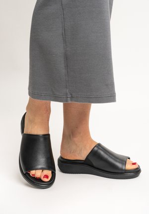 Füße in schwarzen Ledersandalen mit offener Spitze und dicken Sohlen, kombiniert mit grauen gekürzten Hosen.