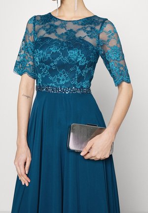 Femme portant une robe en dentelle turquoise avec des manches transparentes et une taille perlée, tenant une petite pochette argentée striée.