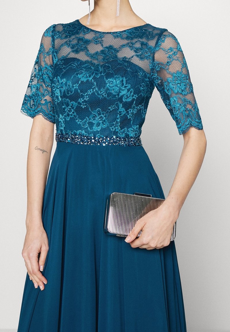 Femme portant une robe en dentelle turquoise avec des manches transparentes et une taille perlée, tenant une petite pochette argentée striée.