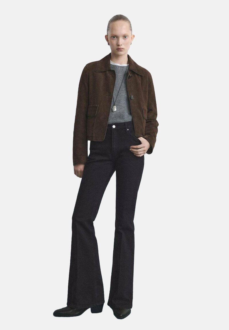 Veste en daim marron, pull en maille grise, et jeans noirs évasés. Quincaillerie simple, tissu texturé, et silhouette ajustée.