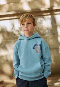 Hellblauer Hoodie mit einer Fronttasche, versehen mit einem "Cool Vibes"-Grafik und einem Smiley-Design in Orange und Dunkelblau. Warmes, glattes Gewebe.