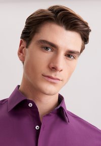 Giovane uomo con capelli castani ben pettinati, indossa una camicia viola a bottone su uno sfondo chiaro e semplice.