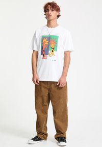 T-shirt blanc en coton avec un design graphique coloré de figures solaires, associé à un pantalon en velours côtelé marron et des baskets noires.