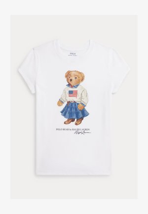 Balta medvilninė marškinėlių su animaciniu lokiu, kuris vilki megztinį su amerikietiška vėliava, džinsinę sijoną ir rudus batus, su tekstu "Polo Bear" žemiau.