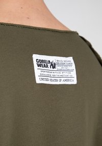 Olijfgroen stof met een gladde textuur heeft een wit label met zwarte tekst waarop "GORILLA WEAR" staat, met details over materiaal en zorginstructies.