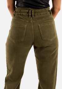 Pantalones de pana verde oliva con cinturilla elástica de talle alto, pliegues en la parte delantera y dos bolsillos traseros. Textura acanalada visible en toda la prenda.