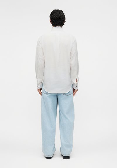 Άνδρας με σγουρά σκούρα μαλλιά που φοράει λευκό μακρυμάνικο πουκάμισο και άνετο ανοιχτό μπλε τζιν, στέκεται γυρισμένος πλάτη σε ένα απλό λευκό φόντο.