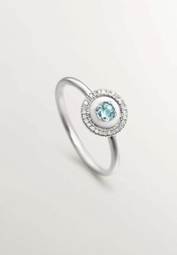 Anillo de plata con un diseño circular, con una piedra central de color azul claro rodeada de pequeñas piedras preciosas transparentes, creando un contraste texturizado.