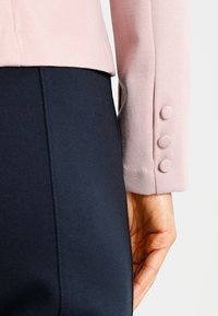 Närbild av en hand bredvid en marinblå kjol och en blommig rosa jackärm med tre matchande tygklädda knappar.