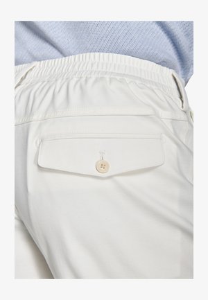 Bílé kalhoty s elastickým pasem a zadní kapsou na knoflík, nošené s texturovaným světle modrým topem.