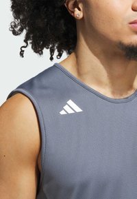 T-shirt de sport sans manches gris avec un tissu texturé, un col rond et un logo blanc sur l'épaule gauche.