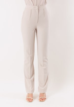 Pantaloni beige a vita alta con gamba dritta indossati con sandali crema open-toe su sfondo bianco.