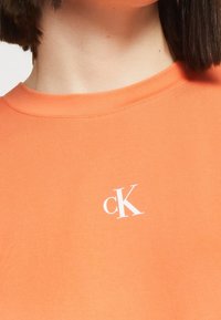 Camiseta de algodón naranja con cuello redondo y logo blanco de "ck" en el lado izquierdo del pecho. Textura suave, corte casual y mangas cortas.