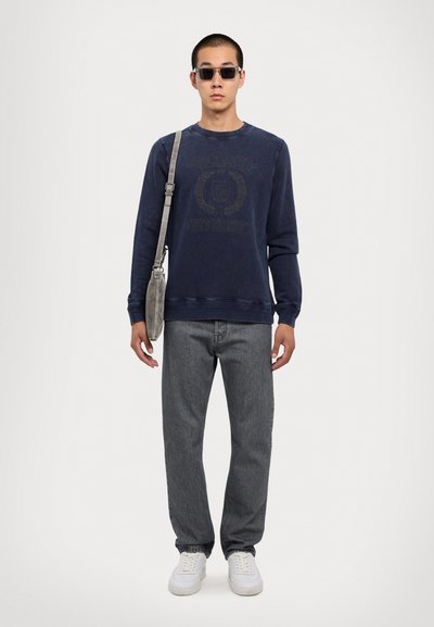 Blå sweatshirt med logo-print, grå jeans, hvide sneakers og en grå crossbody taske. Modellen bærer rektangulære solbriller.