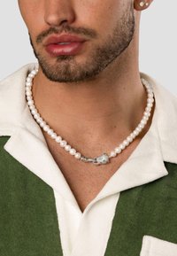 Perlekæde med runde hvide perler, med en sølvklaps lås formet som en hængelås med en lille grøn accent, hvilende på en grøn skjorte med krave.