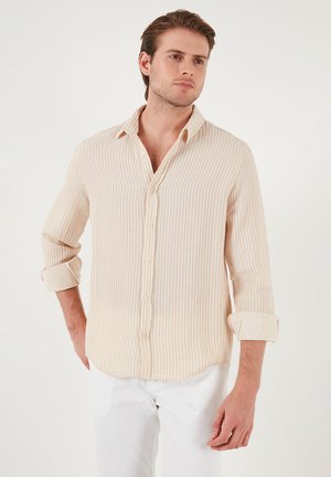Camisa de color beige claro, con botones, rayas horizontales, mangas largas enrolladas hasta los codos y textura de tela suave. Diseño minimalista, estilo casual.
