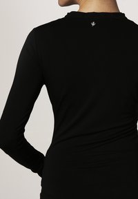 Vrouw in een aansluitend zwart lange mouwen shirt, gezien vanachter met haar linkerarm gebogen en rustend op haar heup.