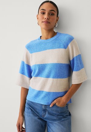 Kvinde iført blå og beige bredstribet kortærmet sweater og blå jeans, stående med den ene hånd i lommen mod en lys væg.