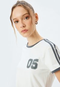 Hvid bomulds T-shirt med grå kant, der har et stort "05" grafik på fronten og grå striber langs ærmerne. Blød tekstur.