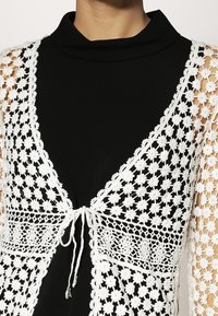 Virkat vitt kofta med blommönster, öppen framsida och snörning; kombinerad med en svart topp med hög krage, vilket visar kontrast i färg.