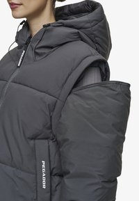 Vadderad väst i mörkgrått med huva, med en framzipper och logodetalj. Tyget ser slätt ut med en något blank yta.