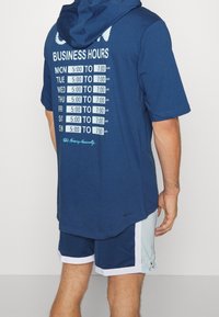 Navyblaues Kapuzen-T-Shirt mit einem aufgedruckten Zeitplan der Öffnungszeiten auf dem Rücken, mit weißem Text und hellblauen Akzenten, kombiniert mit Shorts.