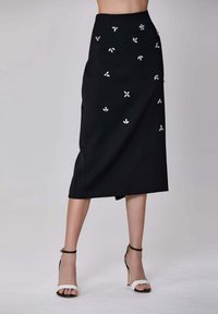 Jupe noire midi avec des embellissements floraux en argent, coupe ajustée, légère fente à l'arrière, texture de tissu lisse, associée à des chaussures à talons noir et blanc.