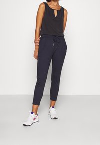 Černý top bez rukávů s výřezem v oblasti klíčové dírky, doplněný o tmavě modré zúžené kalhoty se bočními kapsami a šňůrkou v pase. Oblečeno s bílými teniskami.