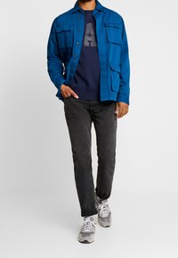 Modrá zapínací bunda s dvěma náprsními kapsami přes námořnický svetr se velkým šedým písmenem. Černé džíny a šedé tenisky dotvářejí celkový vzhled.