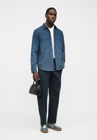 Άνδρας που φοράει μπλε υφασμένη πουκαμίσα με κουμπιά, σκούρο παντελόνι, πράσινα αθλητικά παπούτσια, κρατώντας μικρή μαύρη τσάντα, στέκεται μπροστά σε ανοιχτό γκρι φόντο.