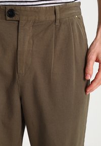 Bruna bomullbyxor med en avslappnad passform, som har frontveck, knappstängning och en jämn yta. Modellens hand vilar på midjan.