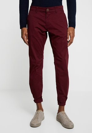 Mężczyzna w podwiniętych bordowych spodniach, ciemnej bluzce z długim rękawem i jasnobeżowych sznurowanych tenisówkach, stojący na jasnej podłodze.