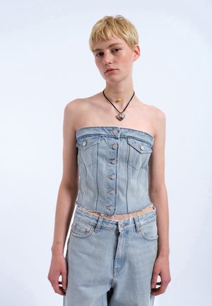 Ung person med kort blondt hår iført stropløs denim korstetop og matchende denim jeans, accessoriseret med hjertevedhæng og gul halskæde.