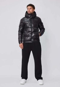Veste matelassée noire avec col montant, finition brillante, fermeture éclair et poignets élastiques, associée à un pantalon noir et des baskets blanches.