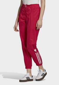 Osoba v jasně červených teplákových kalhotách Adidas s bílými pruhy a logem, ve spojení s bílými teniskami Adidas s černými pruhy.