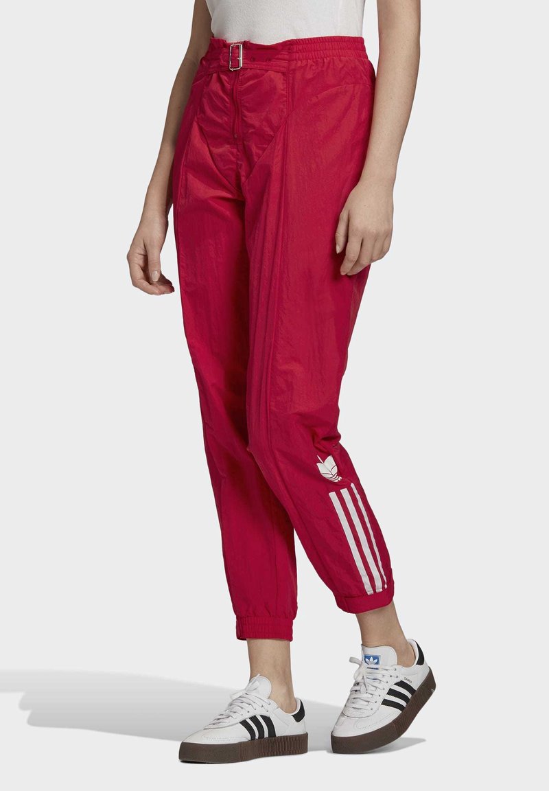 Osoba v jasně červených teplákových kalhotách Adidas s bílými pruhy a logem, ve spojení s bílými teniskami Adidas s černými pruhy.