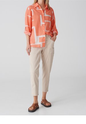Mujer con camisa coral suelta con botones y patrones abstractos blancos, pantalones beige recortados y sandalias marrones con suelas negras.