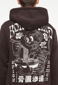 Donkerbruine hoodie met een grote, grafische print van een skelet dat ramen eet met eetstokjes, gedetailleerd met witte tekst en ontwerpen.