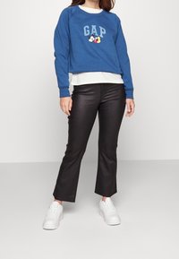 Blå tröja med "GAP"-logotyp och Mickey Mouse-grafik, lager på en vit topp, kombinerat med svarta utsvängda byxor och vita sneakers.