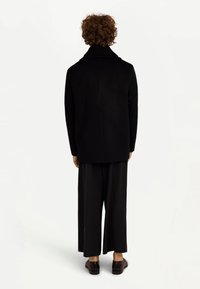 Svart kappa i ullblandning med hög krage, rak skärning och långa ärmar, kombinerad med lösa svarta byxor och mörka slip-on skor.