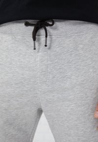 Hellgraue Jogginghose mit schwarzem Kordelzug, der in einer Schleife an der Taille gebunden ist, getragen mit einem schwarzen Oberteil.