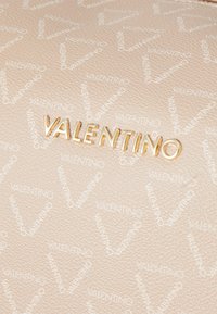 Μπεζ υφασμάτινο δέρμα με επαναλαμβανόμενο λογότυπο, featuring χρυσά ανάγλυφα γράμματα "VALENTINO" που προβάλλονται έντονα.